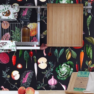 May include: A kitchen with a black background wallpaper featuring a repeating pattern of vegetables, fruits, and other kitchen-related items. A wooden cutting board hangs on a rack above a counter with a set of beeswax food wraps and two red apples on a white plate.