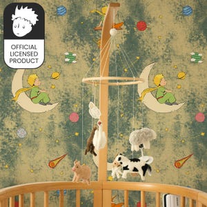 May include: A baby mobile with felt animal figures, including a cow, sheep, and pig, suspended from a wooden frame. The mobile hangs over a wooden cot. The background is wallpaper with illustrations of the Little Prince.
