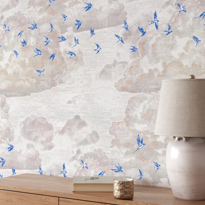 Pode incluir: Um papel de parede branco com um padr&atilde;o texturizado sutil que apresenta um desenho repetitivo de p&aacute;ssaros azuis voando entre nuvens marrons claras. Uma l&acirc;mpada com um abajur branco e uma base de cer&acirc;mica branca est&aacute; sobre uma mesa de madeira com um livro e um objeto decorativo.