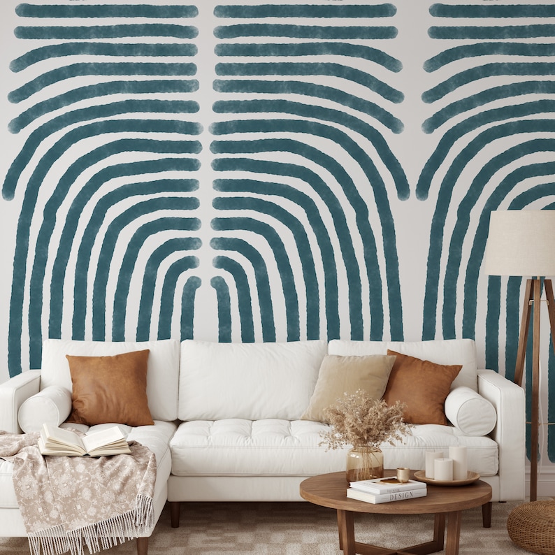 May include: A white couch with brown and beige throw pillows sits in front of a wall with a teal blue abstract pattern. The couch is in a living room setting with a wooden coffee table and a lamp.