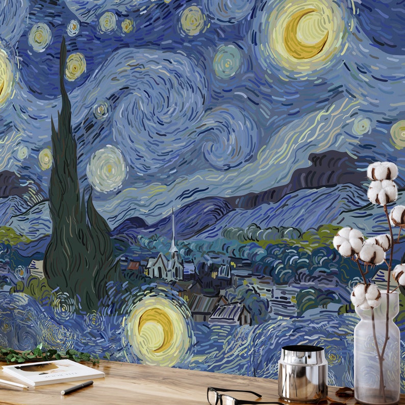 May include: A blue and yellow starry night painting with a town in the foreground. The painting is on a wall with a vase of cotton flowers in front of it.
