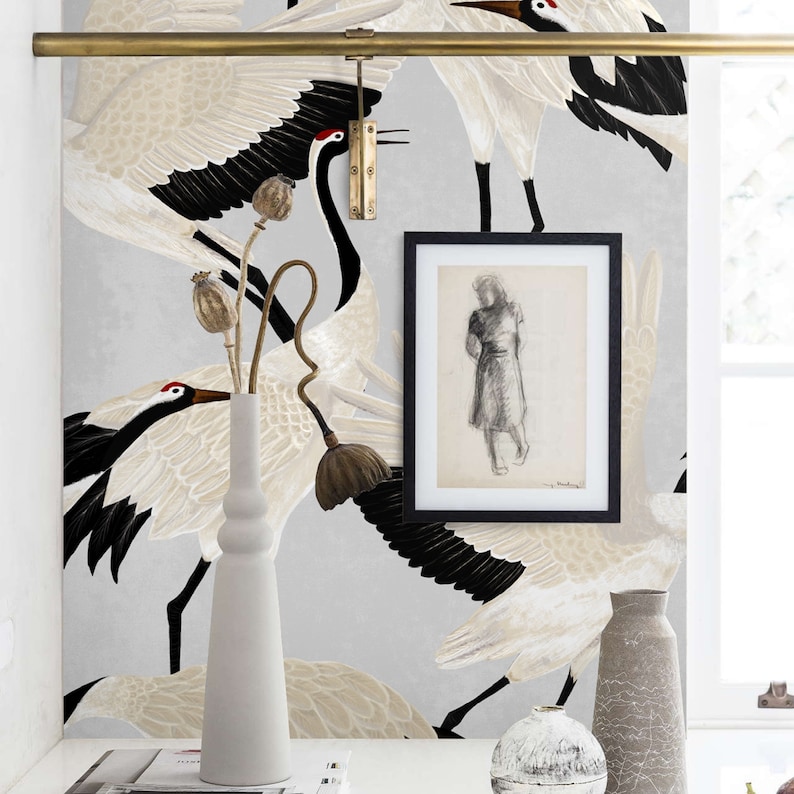 May include: A room interior with a grey crane wallpaper featuring black and white birds. A white vase holds dried stems. A framed sketch hangs on the wall. A brass rod and decorative vases are also visible.