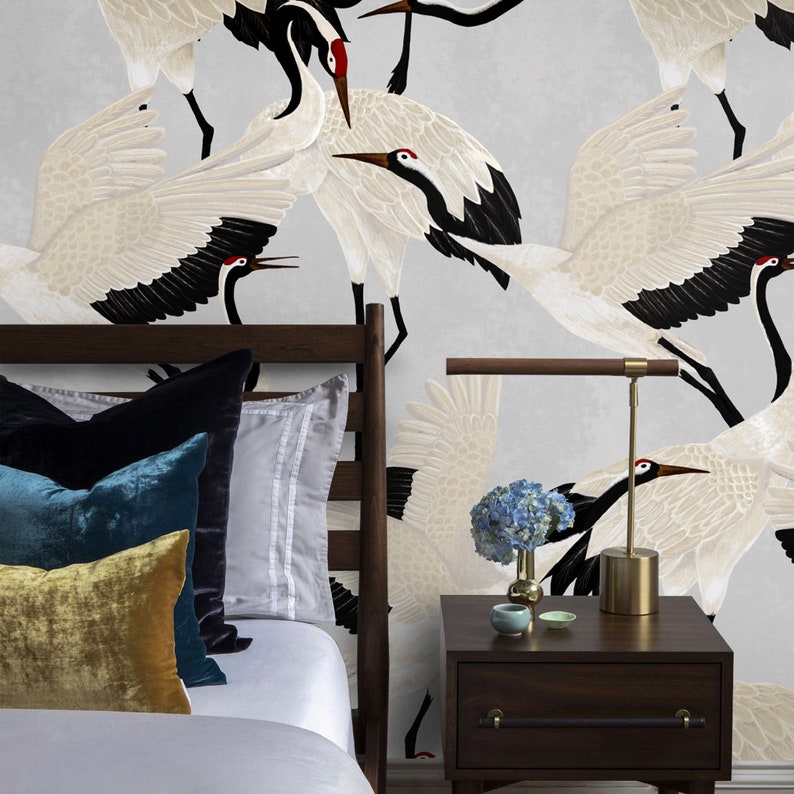 May include: A bedroom scene with a bed featuring velvet cushions in gold, teal, and black. The dark wood headboard is set against wallpaper with a crane pattern. A bedside table holds a lamp and vase with blue flowers.