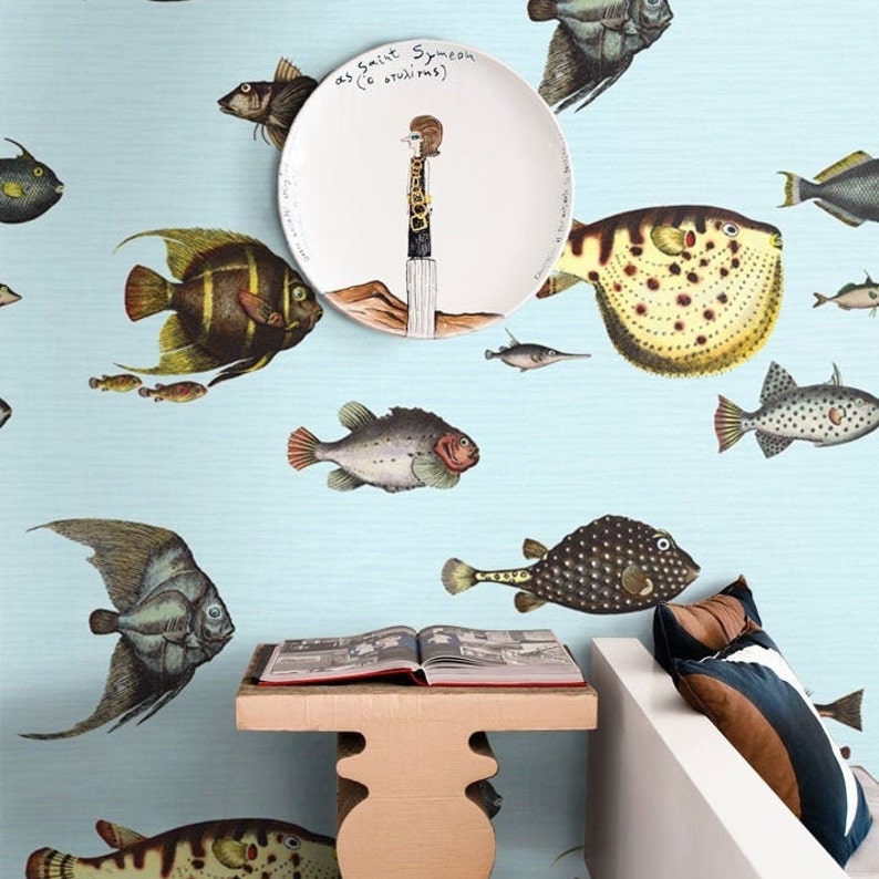 Pu&ograve; includere: Un muro azzurro chiaro con un piatto bianco che presenta un'illustrazione di un personaggio dei cartoni animati in piedi su una roccia. Il piatto &egrave; circondato da un motivo di pesci colorati. Un libro &egrave; su un tavolo di legno davanti a un divano bianco con un cuscino marrone e blu.
