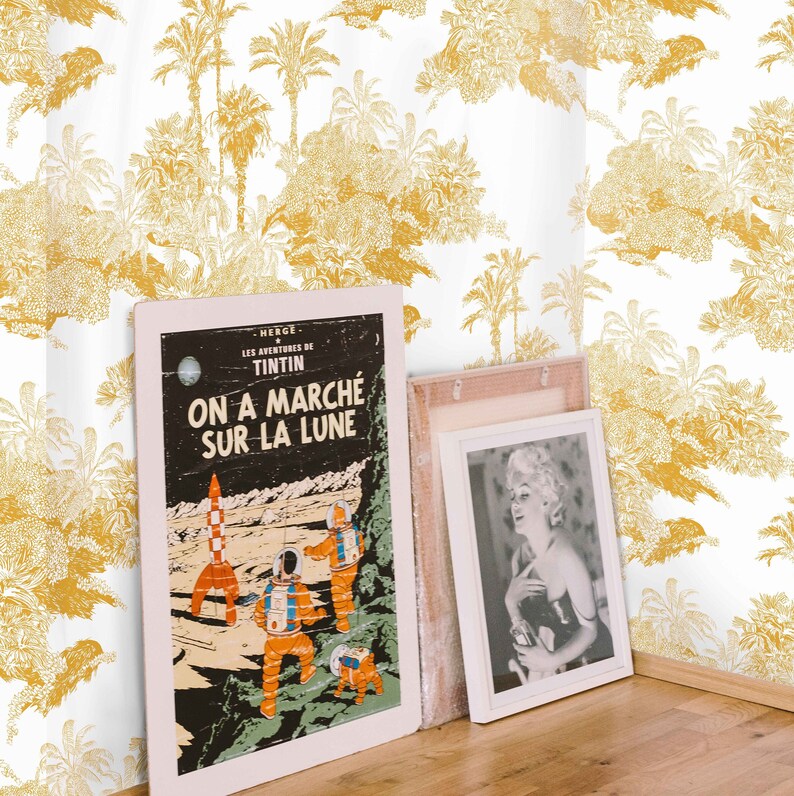 May include: A white wall with a yellow and white palm tree wallpaper pattern. Two framed pictures are leaning against the wall. The picture on the left is a vintage comic book cover with the text "On a march&eacute; sur la lune". The picture on the right is a black and white photo of a woman.