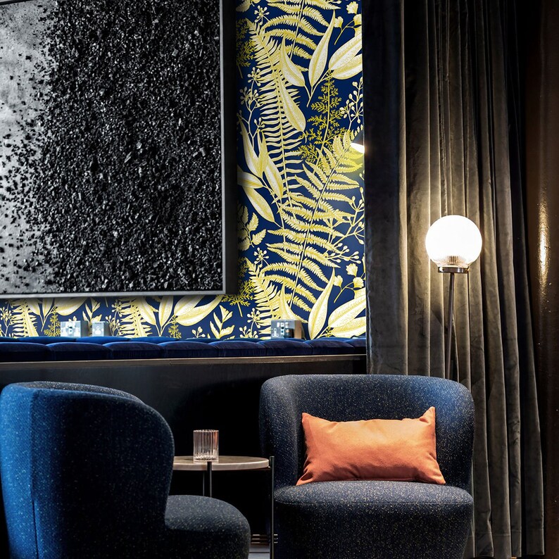 May include: Two blue armchairs with orange throw pillows in a living room with a dark blue and gold floral wallpaper, a black and white painting, and a floor lamp with a white globe.