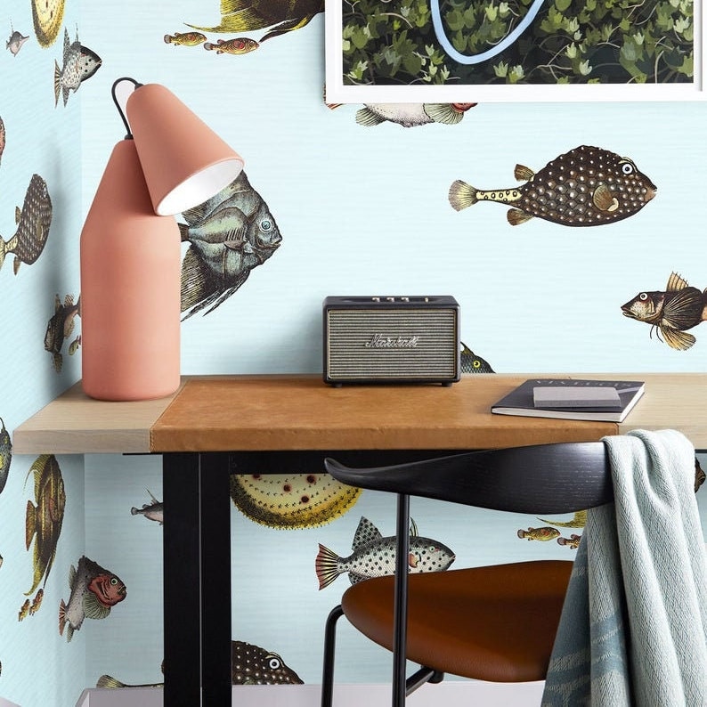 Pu&ograve; includere: Una carta da parati blu chiaro con un motivo a pesce. Una scrivania con un piano in pelle marrone e gambe in metallo nero. Un altoparlante nero e argento con la scritta "Marshall" su di esso. Una sedia nera con una seduta in pelle marrone. Una lampada con una base rosa e un paralume bianco. Una coperta di lana blu e bianca &egrave; drappeggiata sullo schienale della sedia.