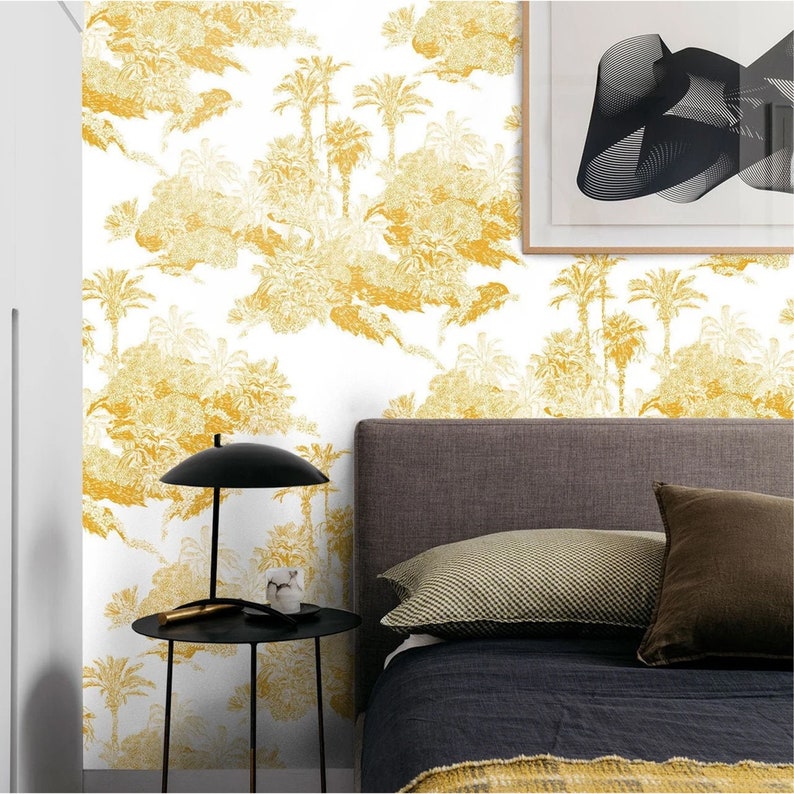 May include: A bedroom with a white wall featuring a yellow palm tree wallpaper pattern. A gray headboard with a black bedspread and pillows is in the foreground. A black side table with a lamp and a small object is next to the bed.
