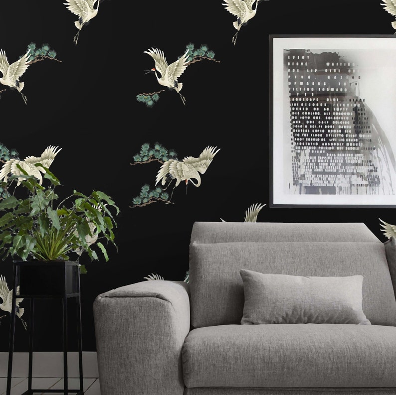 May include: Black and white wallpaper with a repeating pattern of white cranes flying in front of green pine branches. A gray sofa with a gray pillow is in the foreground. A black and white framed print is hanging on the wall.