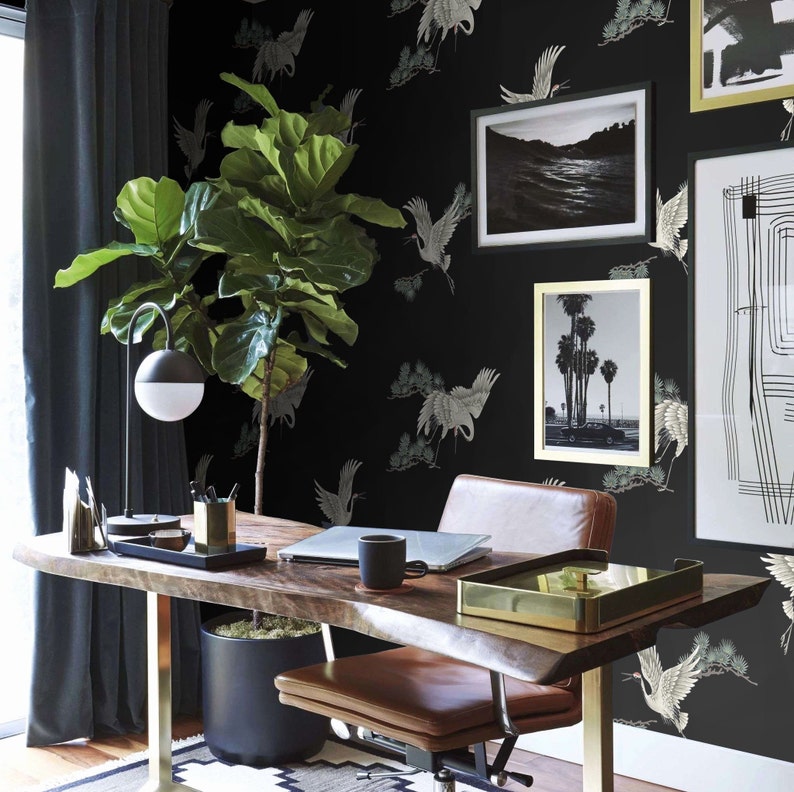 May include: A home office workspace with a black desk and a brown leather office chair. The wall is covered in black wallpaper with a white crane pattern. A large potted plant sits on the desk next to a lamp and a gold tray.