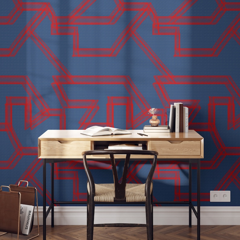 May include: A wooden desk with a black metal frame and a chair in front of it. The wall behind the desk is covered in a blue wallpaper with a red geometric pattern. There are books and a vase with flowers on the desk.