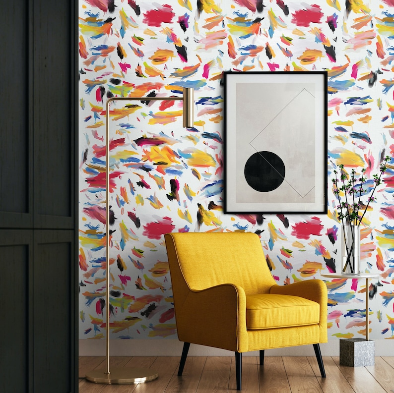 May include: A yellow armchair sits in front of a wall with a colorful abstract pattern wallpaper. A black framed picture with a black circle in the center hangs on the wall. A gold floor lamp stands next to the chair.