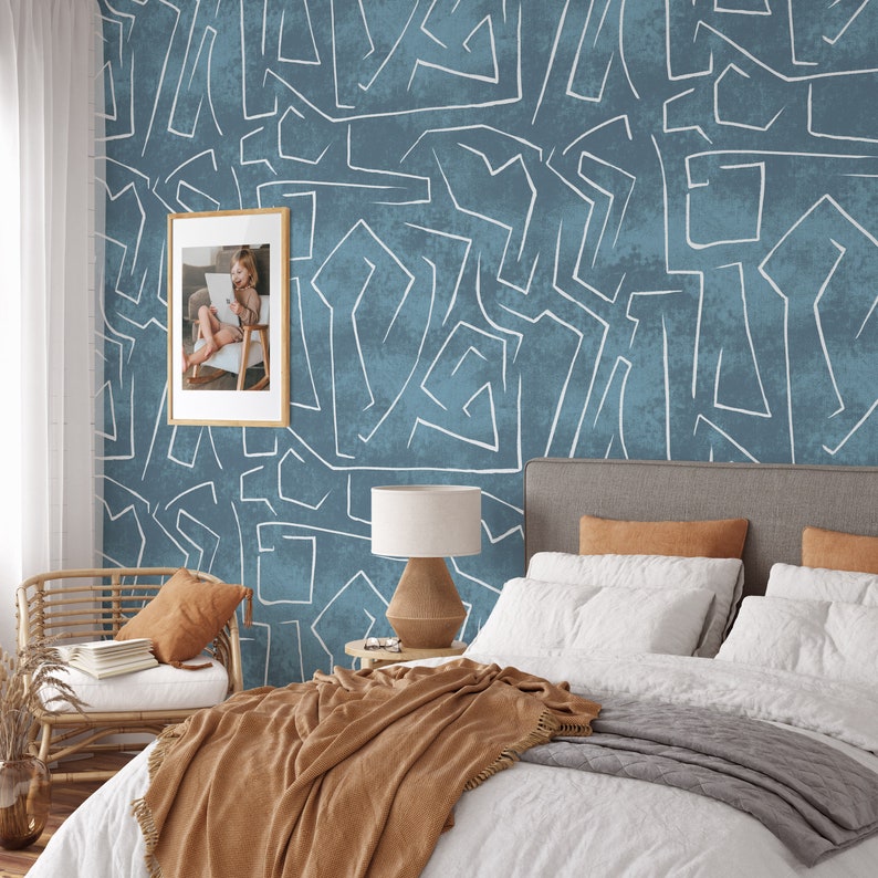 Puede incluir: Un dormitorio con una cama con ropa de cama blanca y una manta marr&oacute;n. La pared est&aacute; decorada con papel pintado azul con l&iacute;neas abstractas blancas. Una foto enmarcada cuelga de la pared y una silla de mimbre est&aacute; en la esquina.