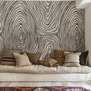 May include: A cozy living space featuring a neutral-toned daybed adorned with various throw pillows. The pillows showcase a range of textures and colors, from woven stripes to solid earth tones. The backdrop is a large wall hanging with a black and white tree ring design.