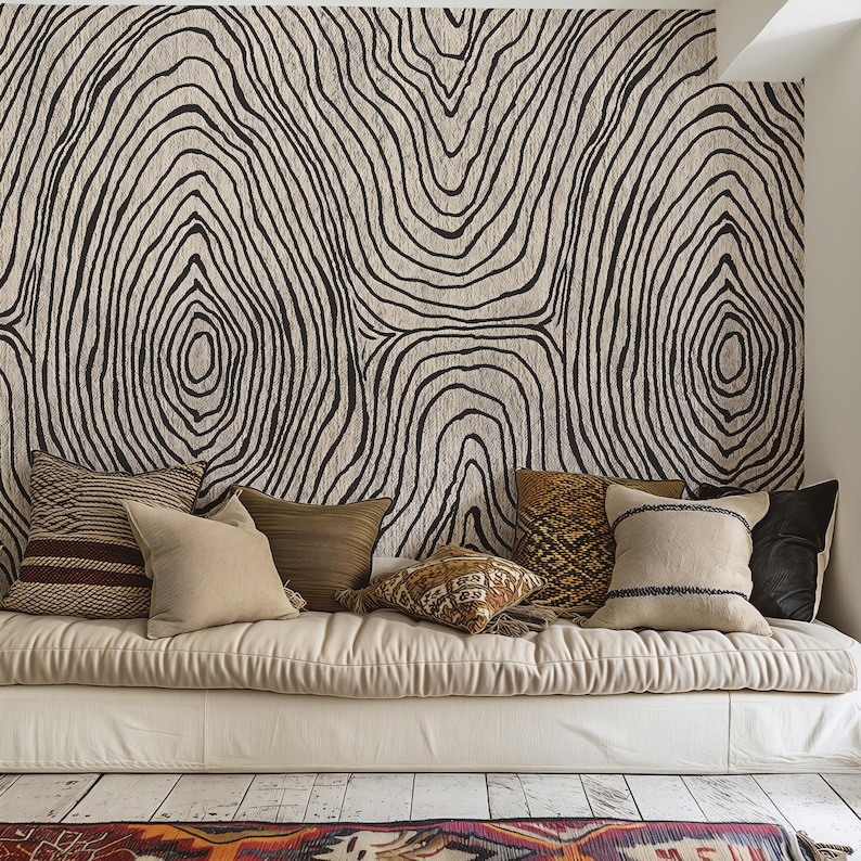 May include: A cozy living space featuring a neutral-toned daybed adorned with various throw pillows. The pillows showcase a range of textures and colors, from woven stripes to solid earth tones. The backdrop is a large wall hanging with a black and white tree ring design.