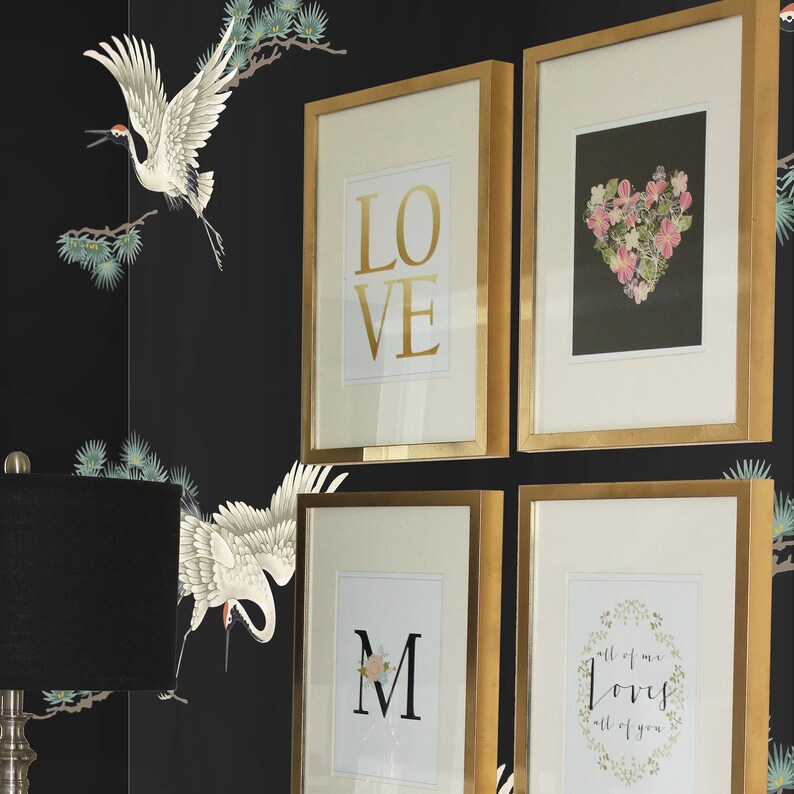 May include: Four gold framed pictures on a black wall with a white crane design. The pictures feature the words "LOVE", "M", and "all of me loves all of you" with a floral wreath. The fourth picture features a pink floral heart.
