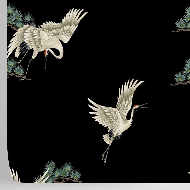 May include: A black background with two white cranes in flight. The cranes have red accents on their heads and are flying over green pine branches.