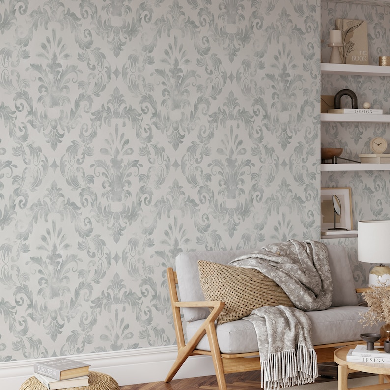 Puede incluir: Un patr&oacute;n de papel pintado damasco gris claro y blanco cubre la pared. Un sof&aacute; gris claro con estructura de madera y un coj&iacute;n tejido est&aacute; frente a la pared. Una manta gris con flecos est&aacute; sobre el sof&aacute;.