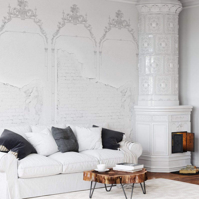 May include: A white sofa with gray pillows sits in front of a white brick wall with a white tiled stove in the corner. The wall has a decorative white pattern on it.