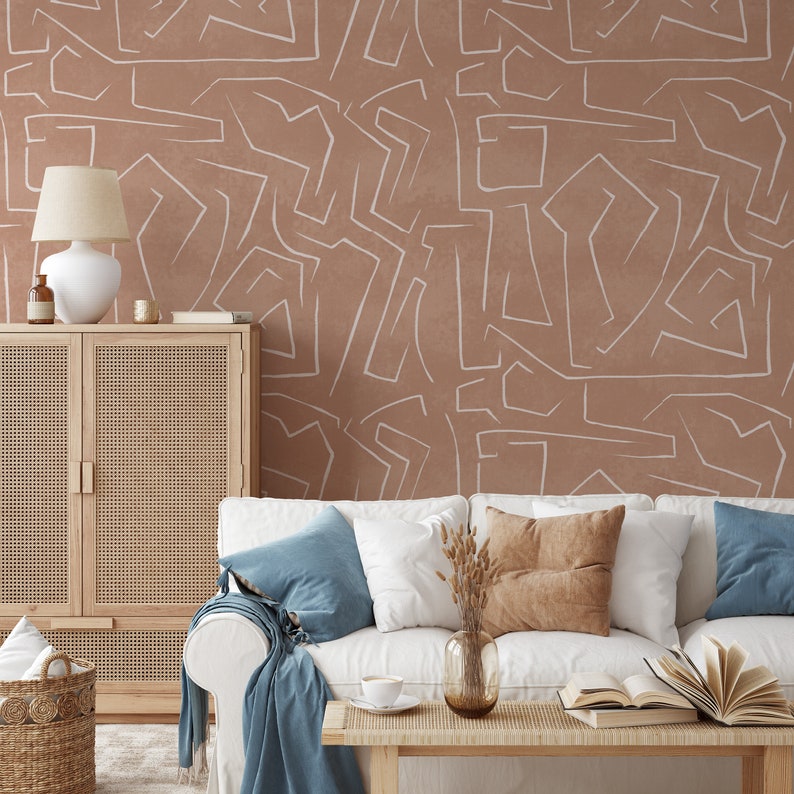 May include: A white couch with blue and brown throw pillows sits in front of a wall with a brown and white abstract pattern. A wooden cabinet with a woven rattan front is to the left of the couch. A vase with dried flowers sits on a coffee table in front of the couch.