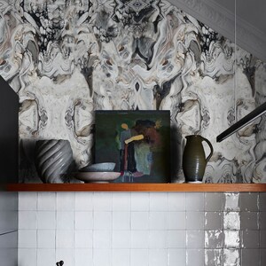 May include: A wooden shelf with a gray and brown abstract patterned wallpaper in the background. The shelf has a gray ceramic vase, a gray ceramic pitcher, a gray ceramic bowl, and a framed painting of a person in a green field.