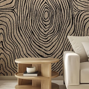 May include: A beige and black abstract pattern resembling tree rings covers a wall. A light wood, two-tiered side table holds a small white bowl and a book. A cream-colored sofa with white pillows is on the right side of the image.