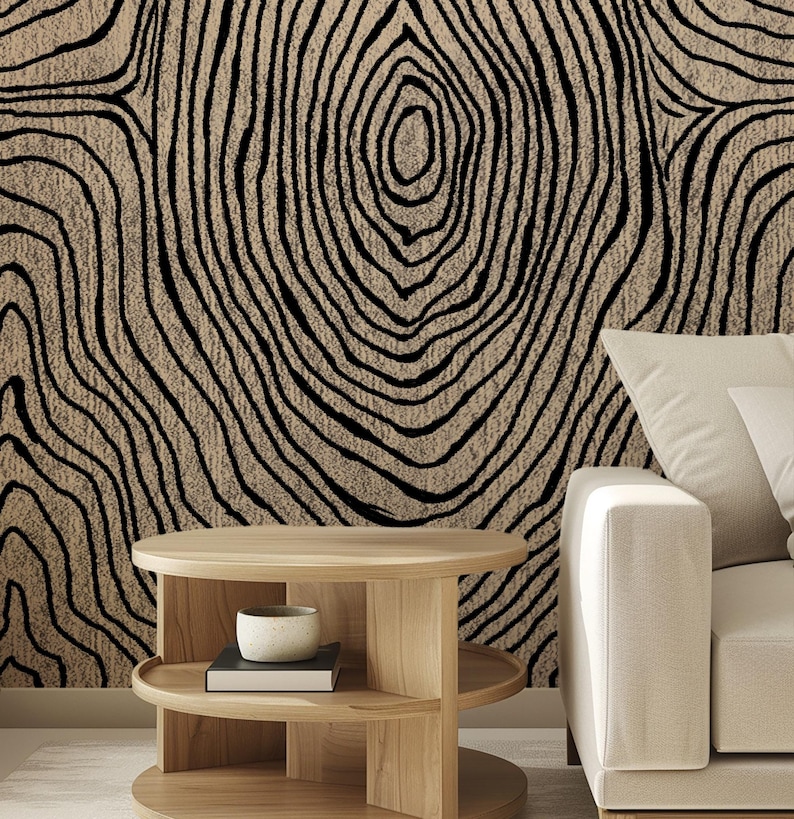 May include: A beige and black abstract pattern resembling tree rings covers a wall. A light wood, two-tiered side table holds a small white bowl and a book. A cream-colored sofa with white pillows is on the right side of the image.
