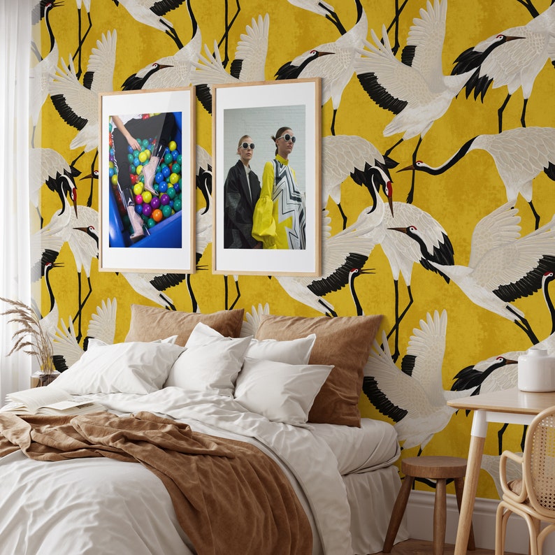 May include: A bedroom with a yellow crane wallpaper and two framed pictures. The wallpaper features a repeating pattern of white and black cranes. The bed has white bedding and brown pillows and a blanket. A small table and chair are in the corner.
