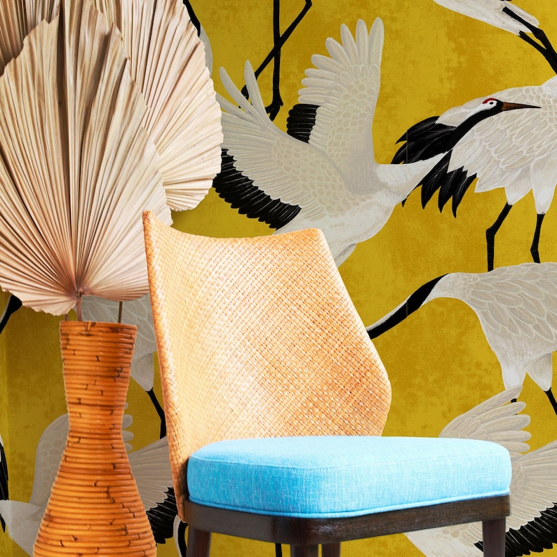 May include: A woven chair with a light blue cushion and a bamboo vase are in front of a yellow background with white and black crane illustrations. A large, dried palm leaf is on the left.