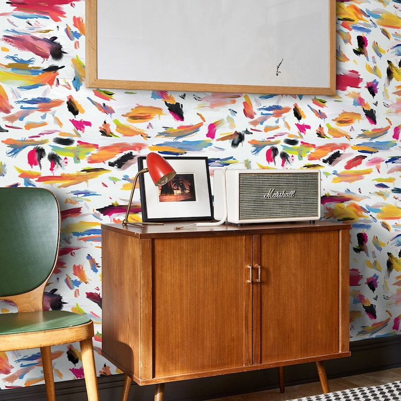 May include: A white wall with a colorful abstract pattern wallpaper. A wooden cabinet with two doors and a white speaker with the word "Marshall" on it. A lamp with a red shade and a framed photo are on top of the cabinet.