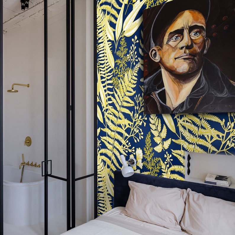 May include: A bedroom with a blue and yellow floral wallpaper featuring ferns. A white bed with a white duvet and a white pillow is in the foreground. A white lamp is on the nightstand. A painting of a man with a black hat is hanging on the wall.