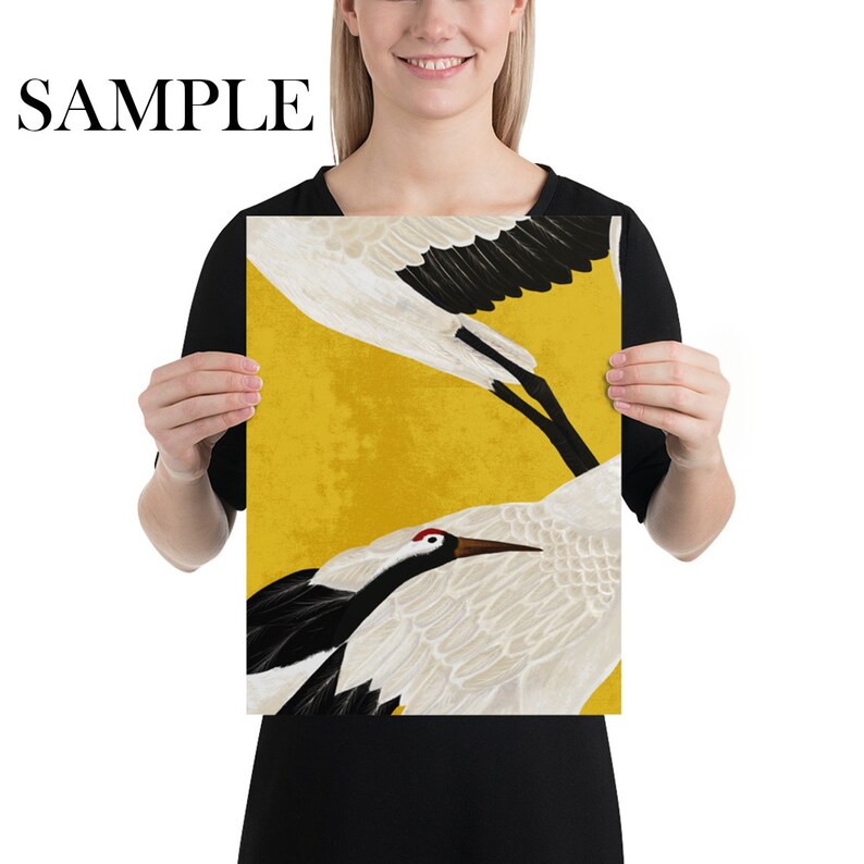 May include: A print featuring two cranes with white feathers, black wings, and red markings on a yellow background. The word "SAMPLE" is printed in black at the top. The artwork is held by a person.