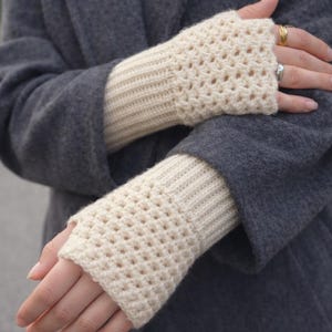 May include: Cream-colored, open-weave fingerless gloves. The gloves have a ribbed cuff and a crocheted top section. The person in the image is wearing a dark gray coat. The gloves are a cozy accessory for cooler weather.