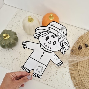 May include: A black and white line art cutout of a scarecrow on a wooden stick, featuring a hat with a leaf detail, stitched patches, and a smiling face. The scarecrow is positioned near one white, one orange, and one olive green pumpkin.