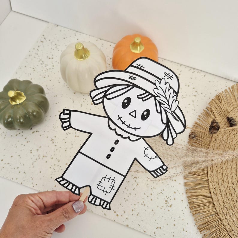 May include: A black and white line art cutout of a scarecrow on a wooden stick, featuring a hat with a leaf detail, stitched patches, and a smiling face. The scarecrow is positioned near one white, one orange, and one olive green pumpkin.