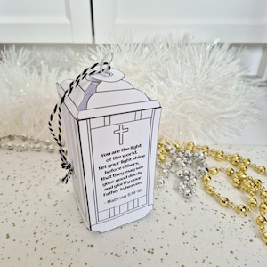 May include: A white paper lantern with a black and white string handle. The lantern features a cross and text from Matthew 5:14-16. Decorative gold and silver beads are in the background.