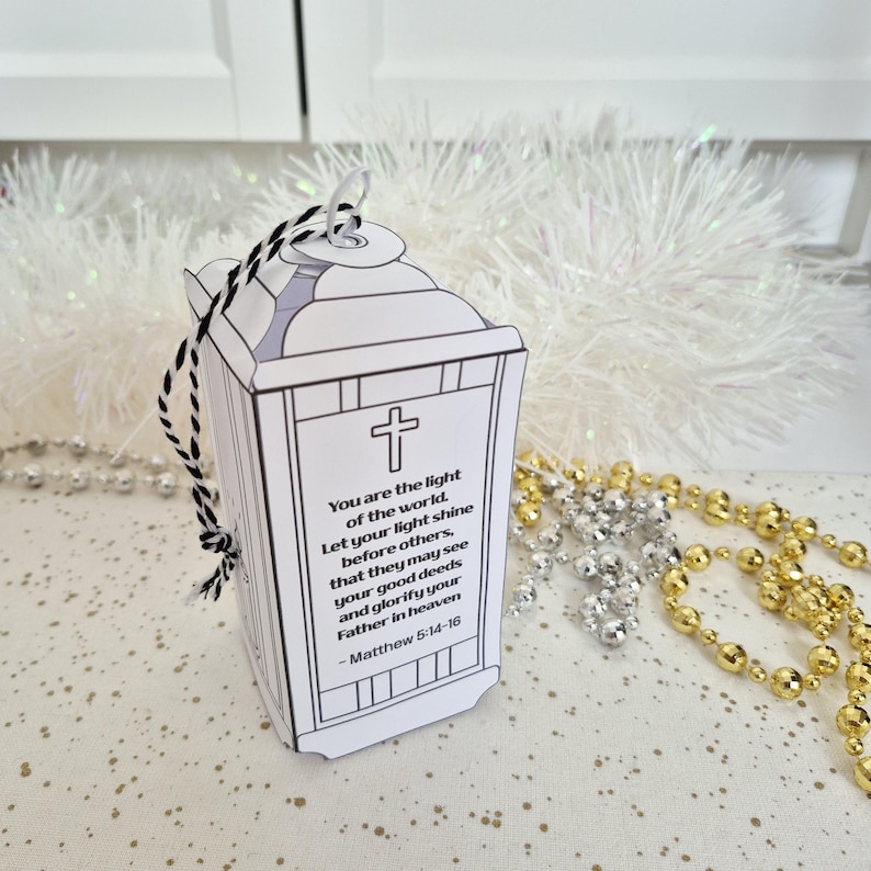 May include: A white paper lantern with a black and white string handle. The lantern features a cross and text from Matthew 5:14-16. Decorative gold and silver beads are in the background.
