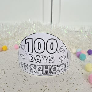 May include: A white paper crown with the text "100 DAYS OF SCHOOL" in black. The crown is decorated with stars and hearts. Colorful pom-poms are scattered around the crown on a white surface.