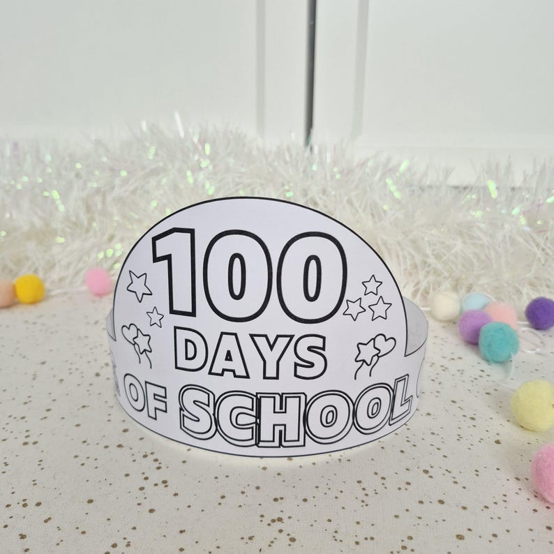 May include: A white paper crown with the text "100 DAYS OF SCHOOL" in black. The crown is decorated with stars and hearts. Colorful pom-poms are scattered around the crown on a white surface.