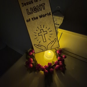 May include: A decorative arrangement featuring a banner with the text "Jesus is the LIGHT of the World" and a cross illustration. A small candle sits on a yellow surface, surrounded by a wreath of red berries.
