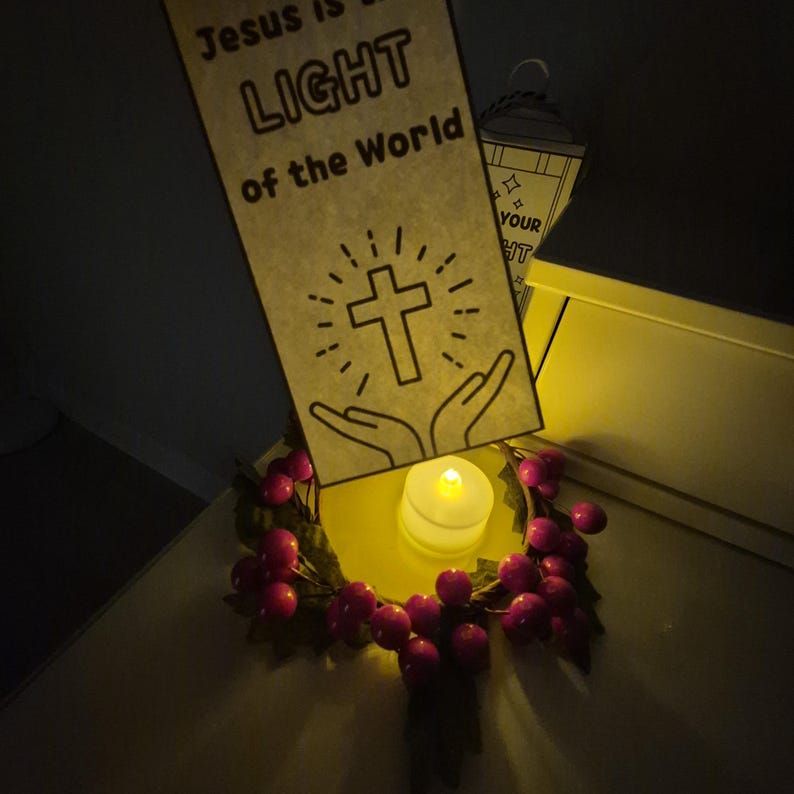 May include: A decorative arrangement featuring a banner with the text "Jesus is the LIGHT of the World" and a cross illustration. A small candle sits on a yellow surface, surrounded by a wreath of red berries.