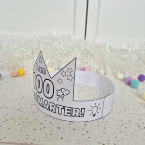 May include: A white paper crown with the text "I am 100% SMARTER!" and star, heart, and lightbulb illustrations. The crown is designed for coloring and celebrating a milestone. The background includes white tinsel and colorful pom-poms.