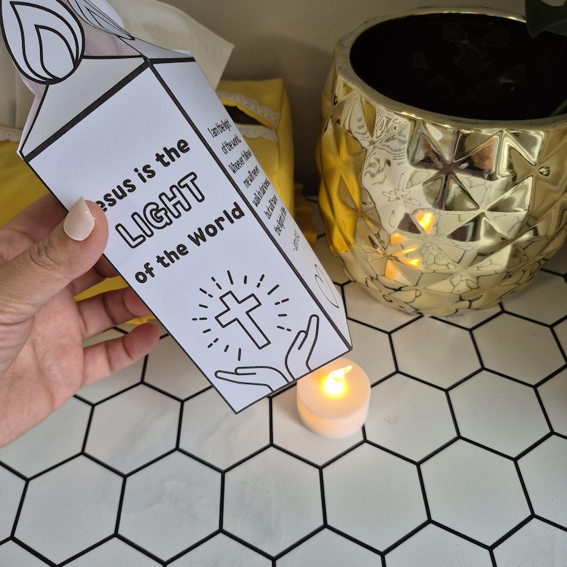 May include: A white paper candle-shaped craft with the text "Jesus is the LIGHT of the World" and a cross illustration. A small, lit, white candle sits below the craft. A gold decorative pot is in the background.