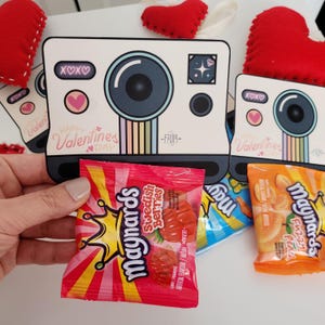 May include: A red and white camera-shaped Valentine's Day card with the text "Happy Valentines Day" and "XOXO" on it. The card is filled with a small bag of red Swedish Berries candy.