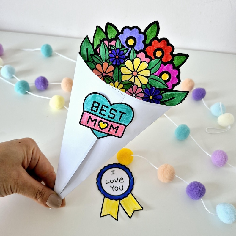May include: A handmade paper bouquet with colorful flowers, held in a white cone. The bouquet has a heart-shaped "BEST MOM" emblem. A "I Love You" ribbon is also present, along with a garland of pastel pom-poms.