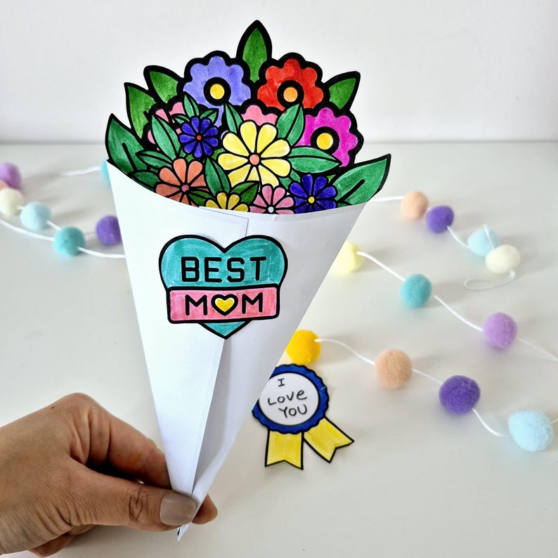 May include: A handmade paper bouquet with colorful flowers and green leaves. The bouquet is in a white cone-shaped holder with a heart-shaped label that says "BEST MOM". A small badge with "I Love You" is also present.