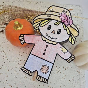 May include: A hand-drawn paper scarecrow cutout featuring a yellow hat with a pink feather, a pink shirt with three black buttons, and blue pants with a brown patch. The scarecrow has a stitched mouth and is positioned next to an orange pumpkin.