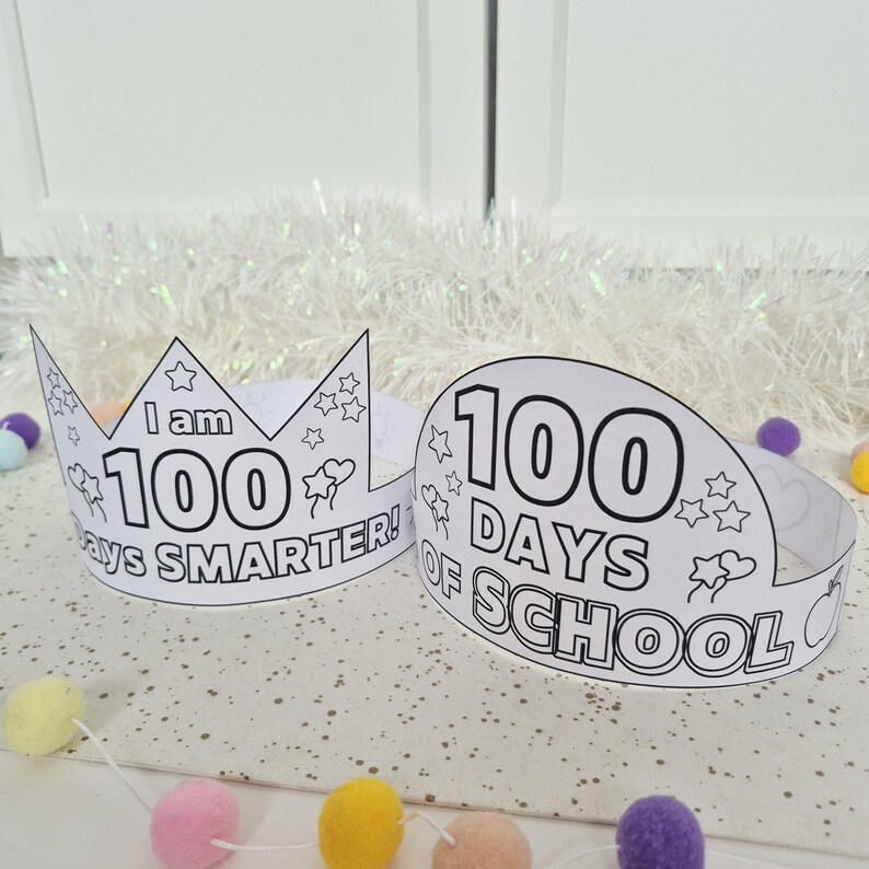 May include: Two white paper crowns with black outlines and text. One crown reads "I am 100 days SMARTER!" and the other reads "100 DAYS OF SCHOOL". Both crowns have star and heart illustrations. The background includes colorful pom-poms.