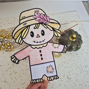 May include: A hand-drawn paper scarecrow puppet on a wooden stick. The scarecrow features a yellow hat with a pink leaf, a peach-colored shirt with three black buttons and a patch, and light blue pants with a brown patch, all outlined in black.