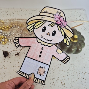 May include: A hand-drawn paper scarecrow puppet featuring a yellow hat with a pink leaf, a peach-colored face with a stitched smile, a pink shirt with three black buttons and a patch, and blue pants with a tan patch, attached to a wooden stick.